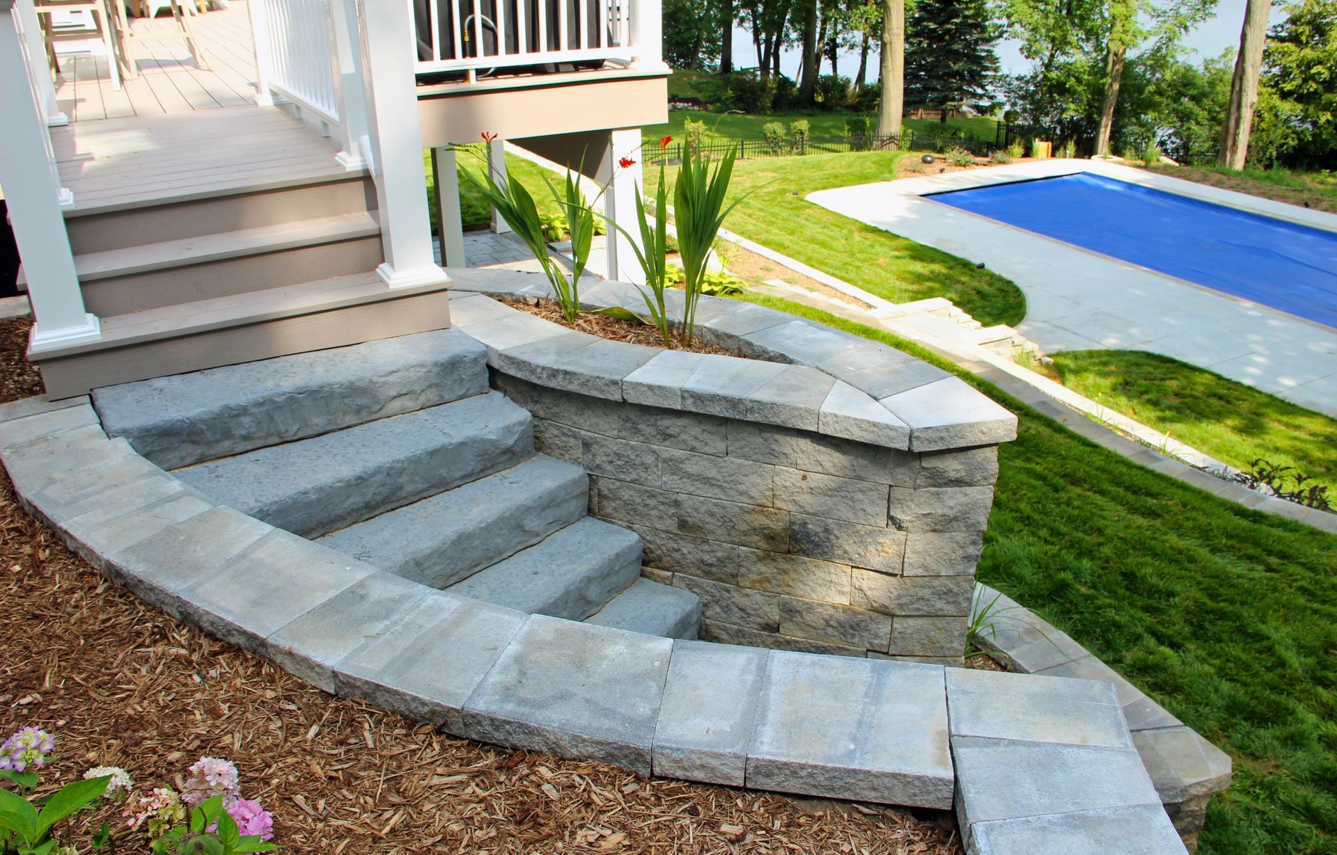 Curved stone steps with retaining wall beside a porch, leading to a landscaped yard and blue pool cover