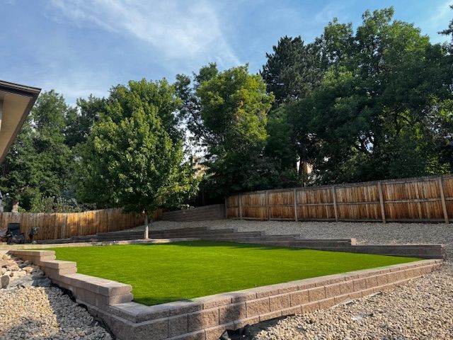 Backyard with a raised green lawn, stone retaining walls, gravel, and a wooden fence under trees.