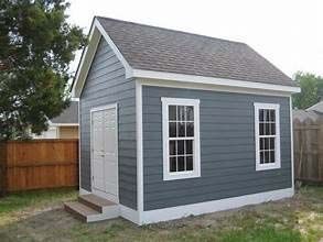 A grey, gabled-roof shed with white trim, two white windows, and double doors with small steps in a grassy backyard.