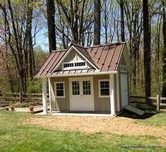 A small tan shed with a dark brown metal roof, white double doors, and a decorative dormer window in a wooded yard.