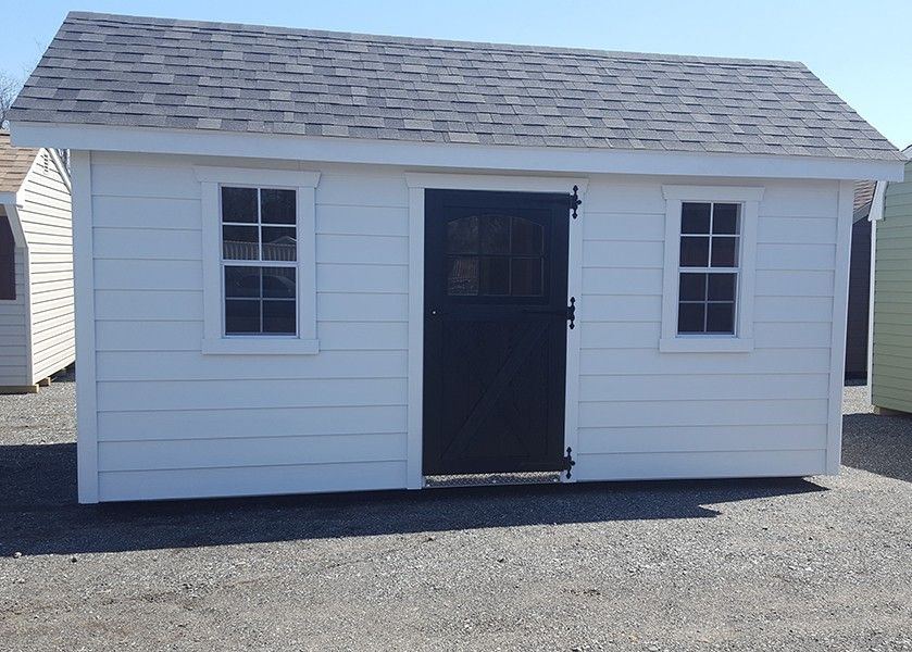A small white storage shed with a black door, shingled roof, and two windows, situated on a gravel surface.