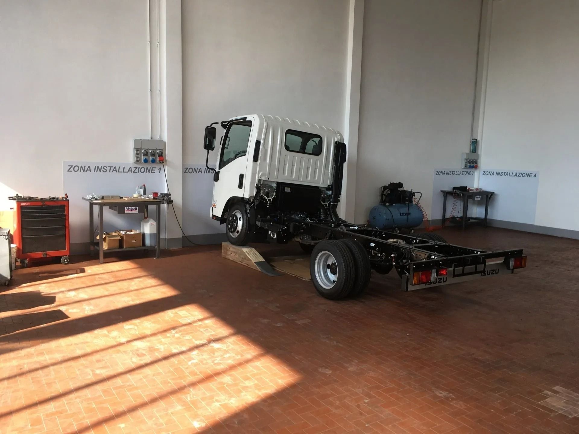 Telaio di camion bianco in un garage industriale con pavimento rosso e attrezzature lungo la parete.