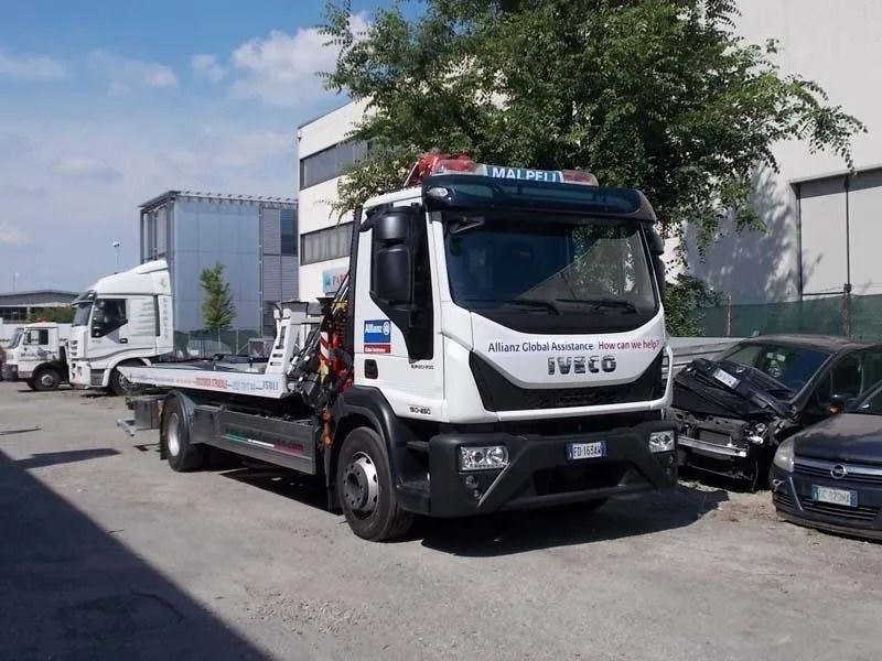 Carro attrezzi bianco con pianale ribassato parcheggiato in un piazzale accanto ad altri veicoli e a un edificio