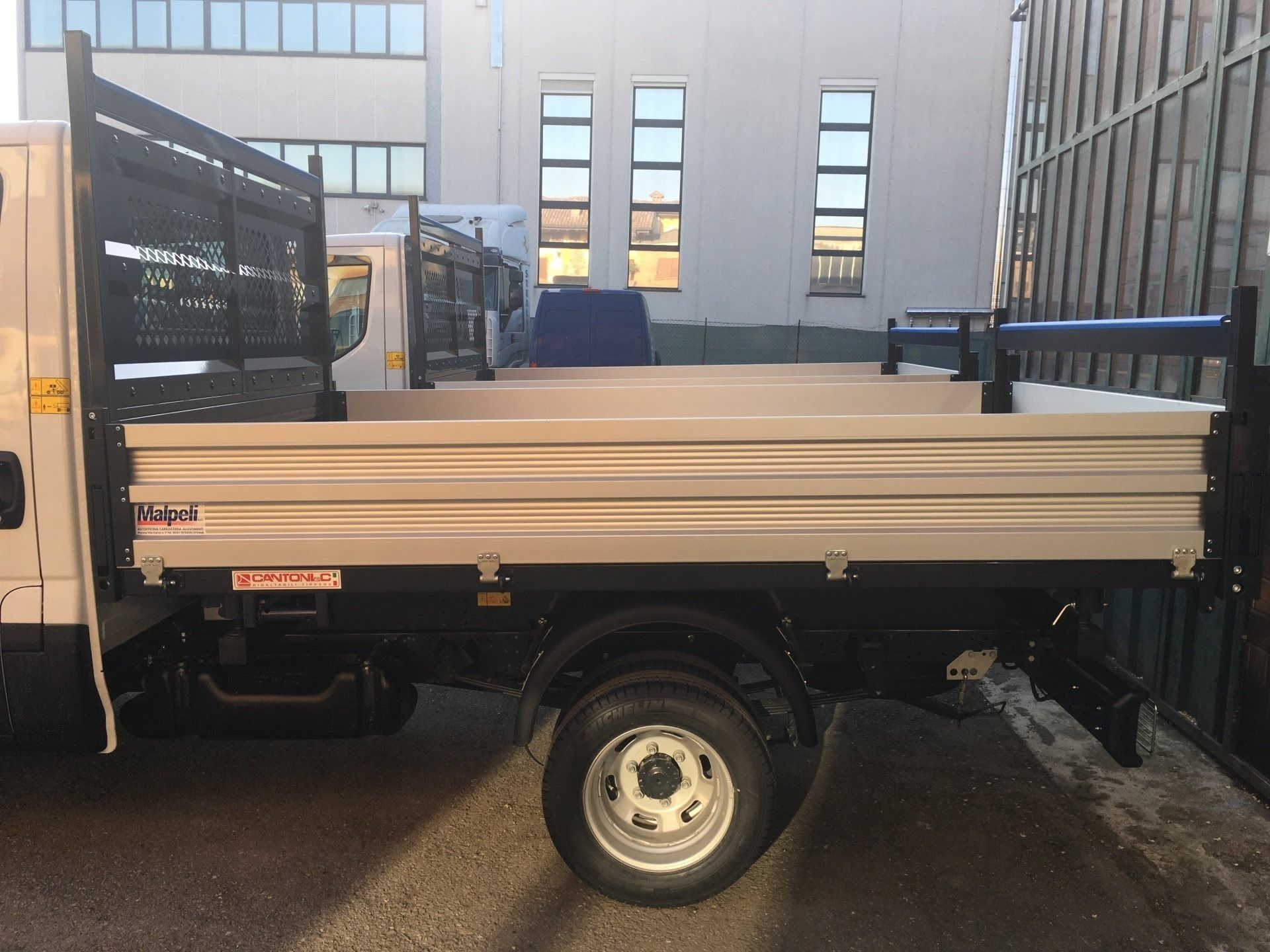 Camion bianco a pianale ribassato parcheggiato in un vicolo accanto a un edificio, vista laterale.