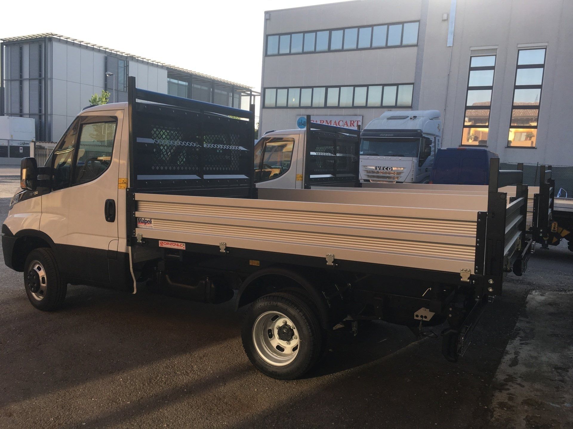 Un camioncino bianco con pianale ribassato parcheggiato davanti a moderni edifici per uffici al crepuscolo.