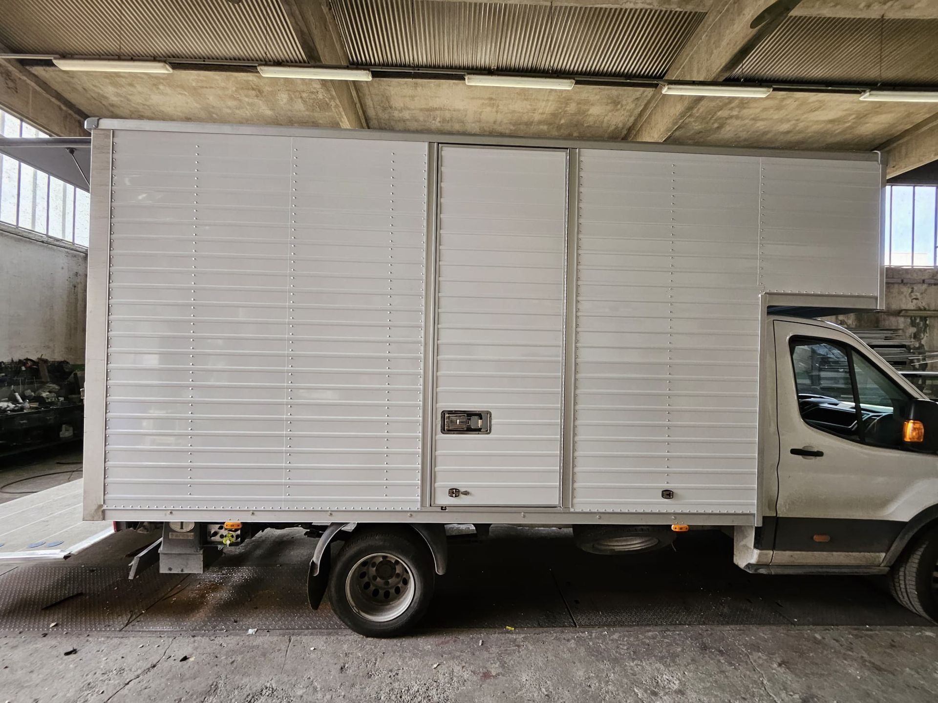 Camion furgonato bianco parcheggiato sotto una tettoia, vista laterale con vano di carico visibile.