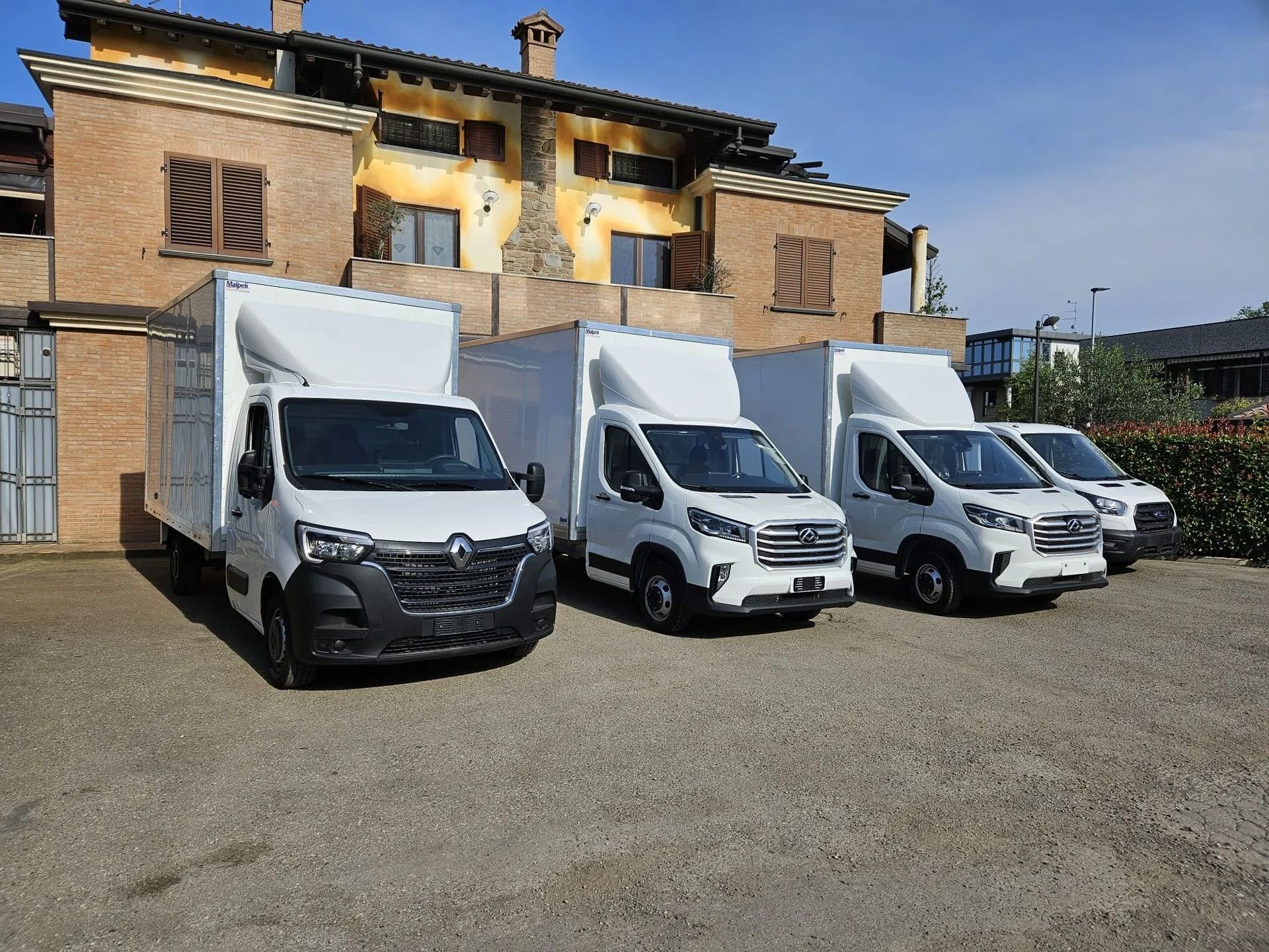 Tre furgoni bianchi per le consegne erano parcheggiati davanti a un edificio rustico su un piazzale ghiaioso.