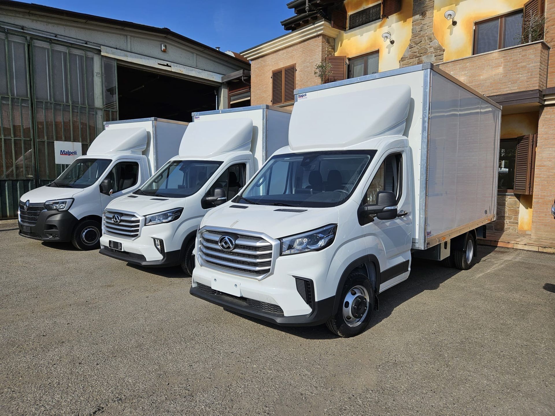 Tre furgoni bianchi parcheggiati in un soleggiato piazzale industriale di fronte a un edificio.
