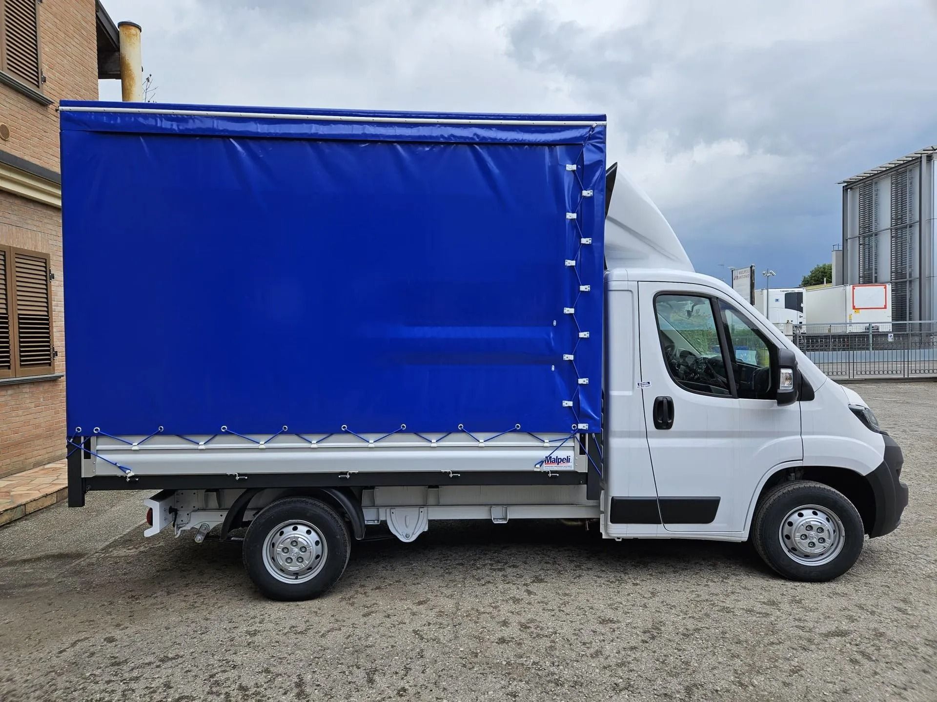 Un furgone bianco con un cassone coperto da un telone blu è parcheggiato accanto a un edificio in mattoni.