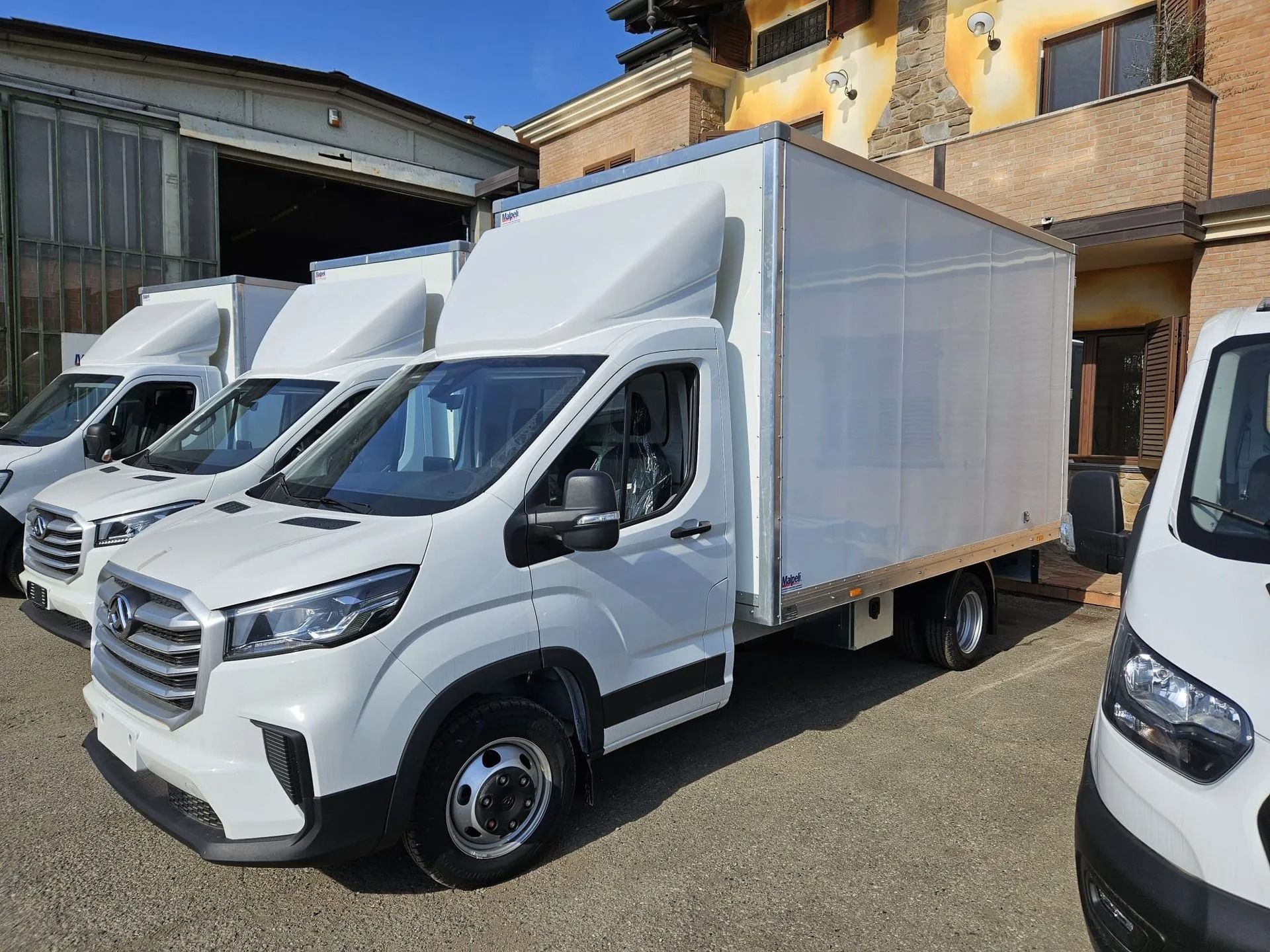 Camion bianchi parcheggiati in fila davanti a un edificio, il camion anteriore inclinato verso la telecamera.