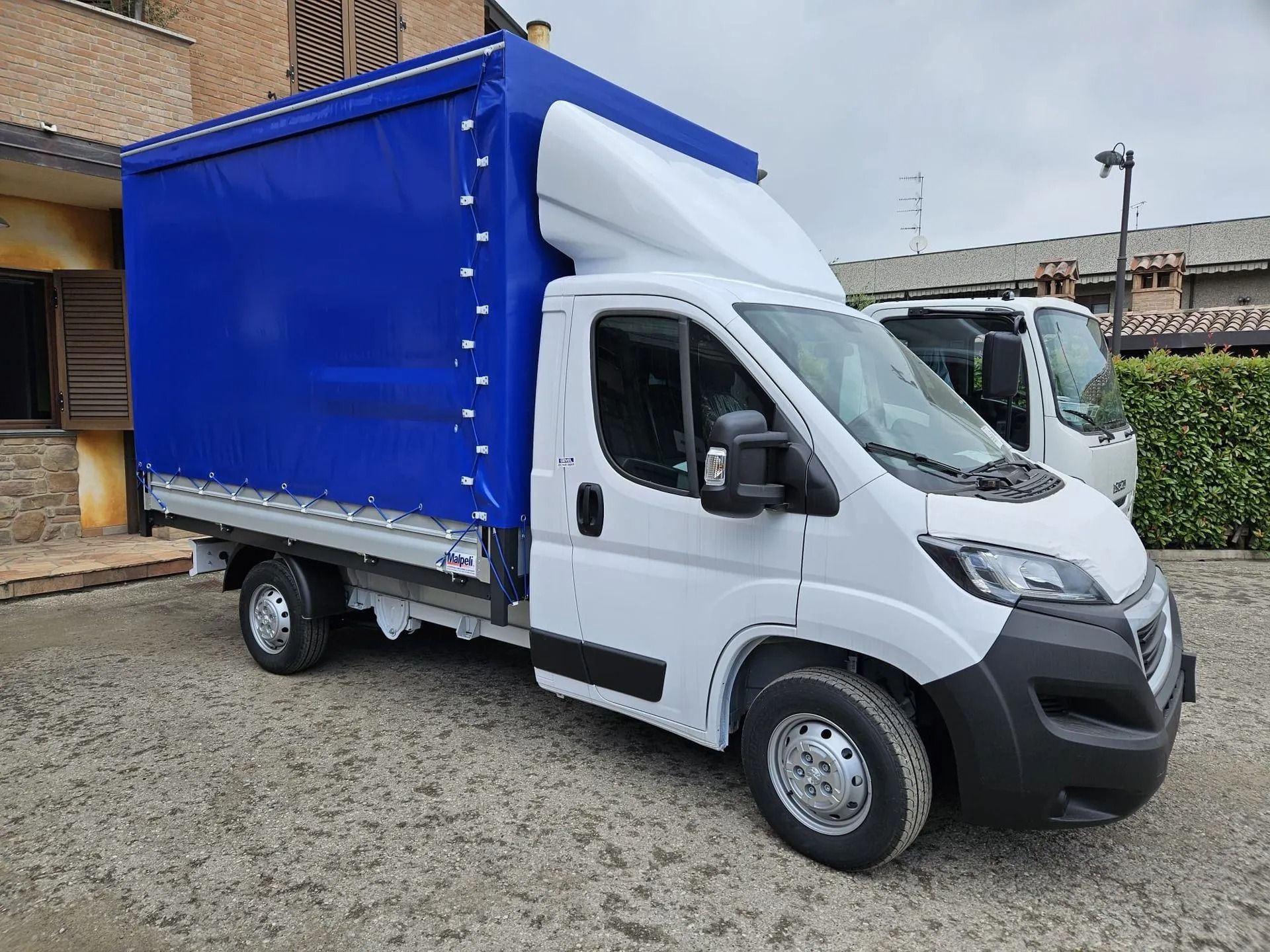 Camion bianco con cabina avanzata e copertura del cassone blu parcheggiato su ghiaia fuori da un edificio