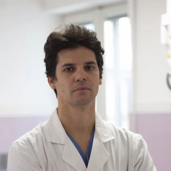 Uomo in camice bianco, guarda dritto negli occhi l'osservatore. Capelli scuri, in un interno.