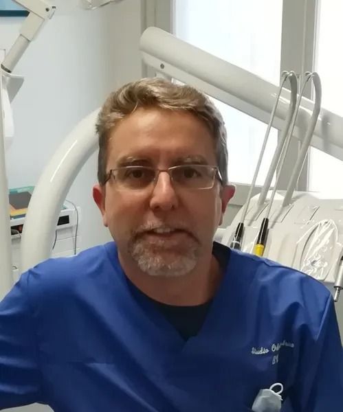 Uomo sorridente in camice blu e occhiali in uno studio dentistico.
