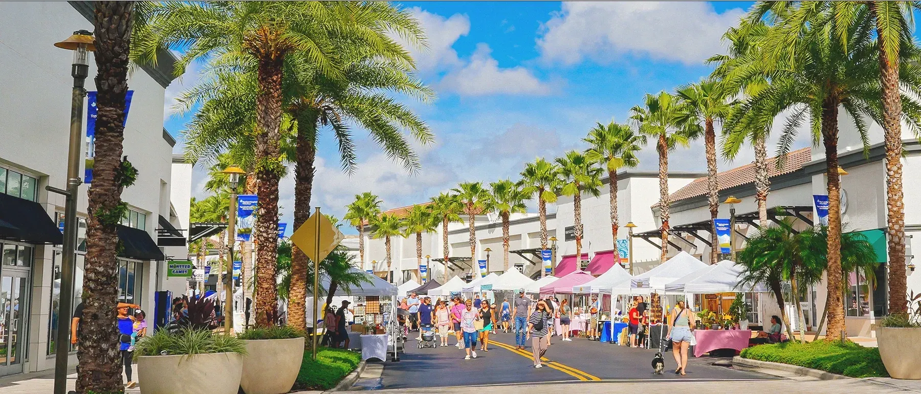 Street market with white tents, palm trees, and people shopping.