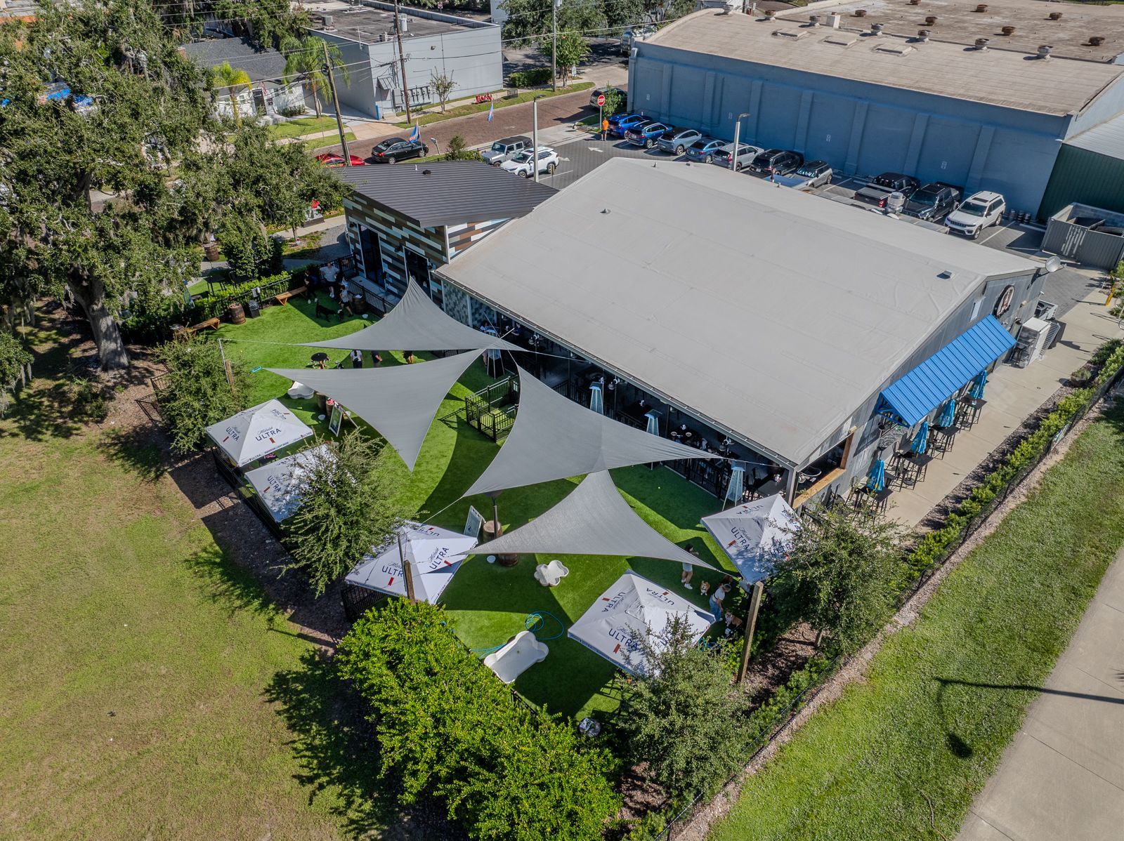 Aerial view of a restaurant with a gray roof and outdoor shaded seating with umbrellas. Green grass surrounds it.