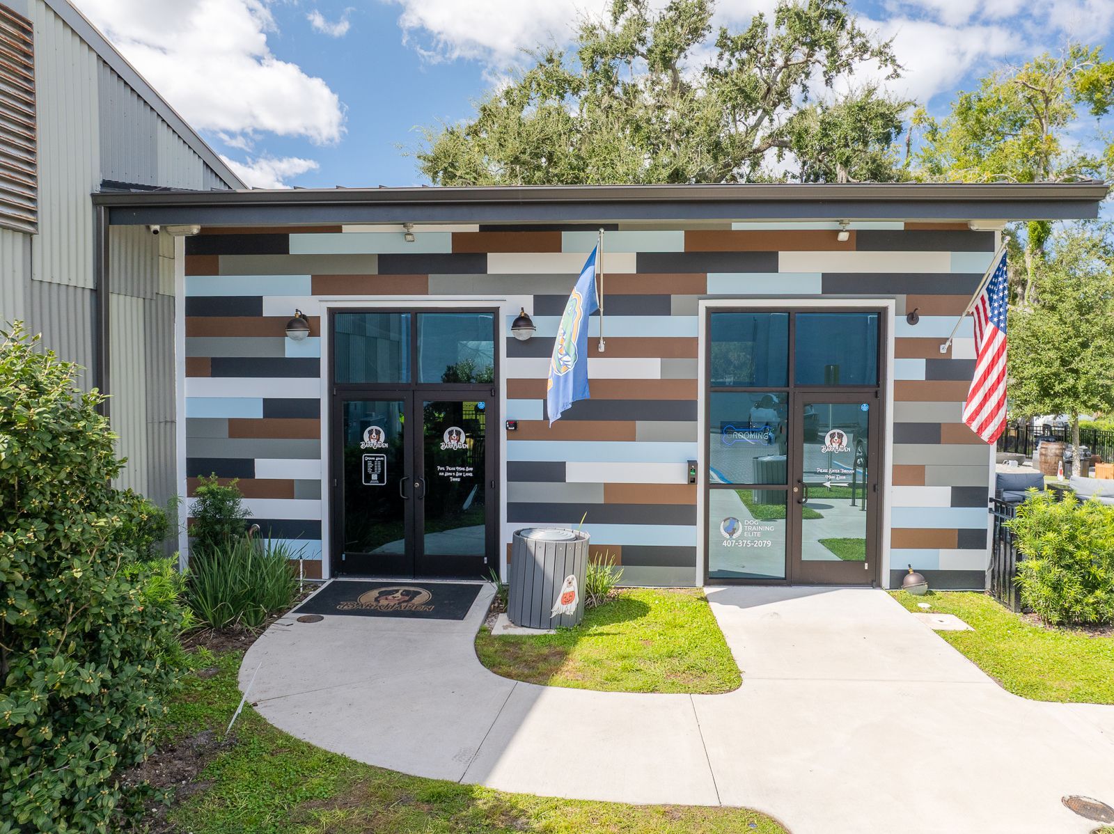 Exterior of a building with two glass doors, striped siding, flags, and a sidewalk.
