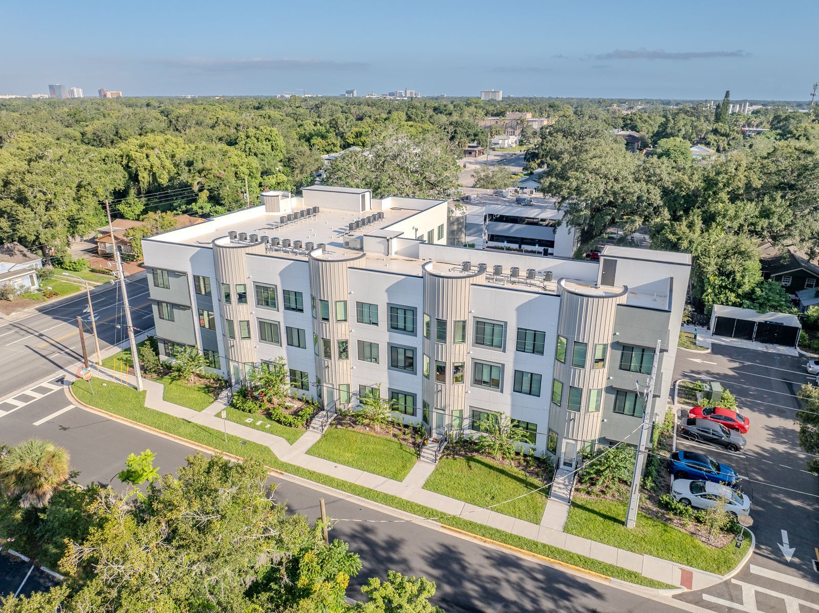 Aerial view of a white multi-story apartment building with green landscaping, trees, and parked cars.