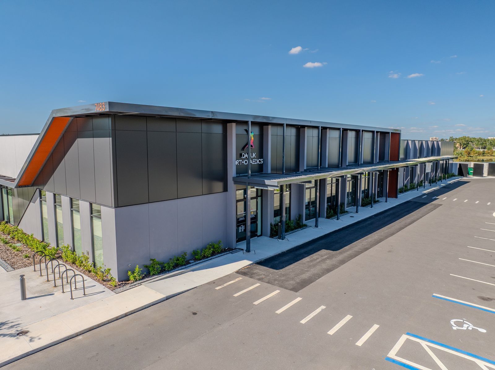 Modern, gray and black building with large windows and a parking lot on a sunny day.
