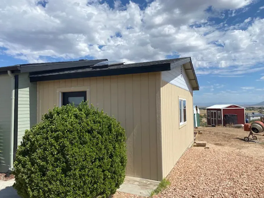 Small beige outbuilding with a green bush in front, gravel yard, and cloudy sky.