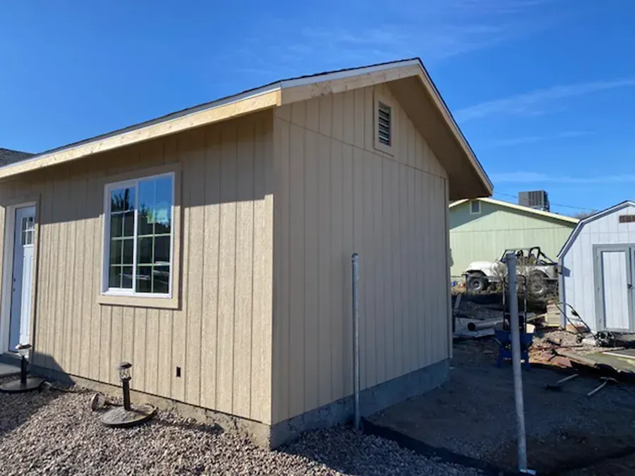 Tan beige shed with a window and white door on gravel, beside a fenced yard under a blue sky