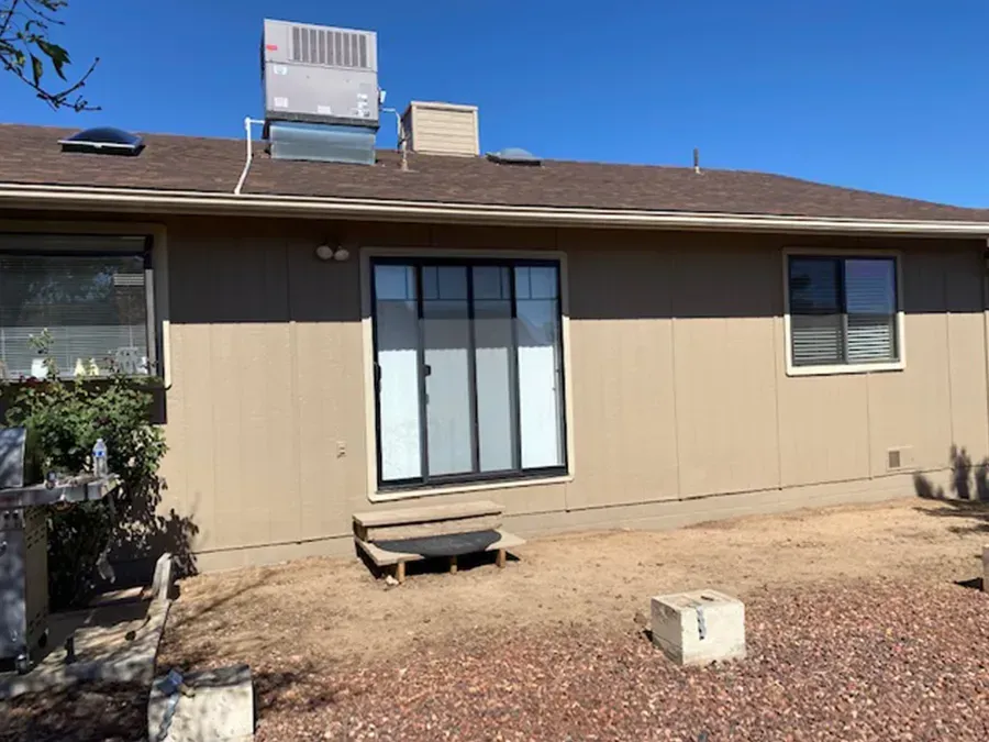 Backyard view of a beige house with sliding glass doors, a small bench, and rooftop HVAC units under a blue sky