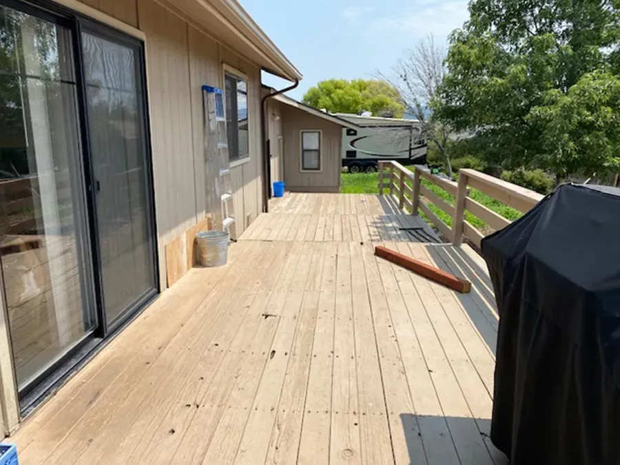 Wooden deck beside a house with sliding glass doors, railing, and a covered grill under a sunny sky