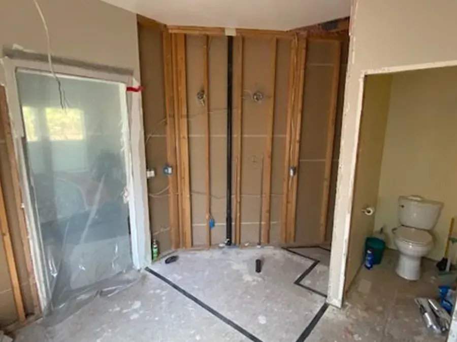 Unfinished bathroom with exposed studs, shower stall at left, toilet at right, and debris on the floor.