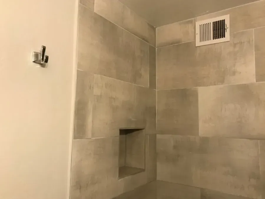 Beige tiled shower corner with recessed niche, wall hook, and vent grate