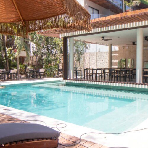 Piscina tropical tipo resort con agua turquesa, palmeras y tumbonas a la sombra bajo un techo de paja.
