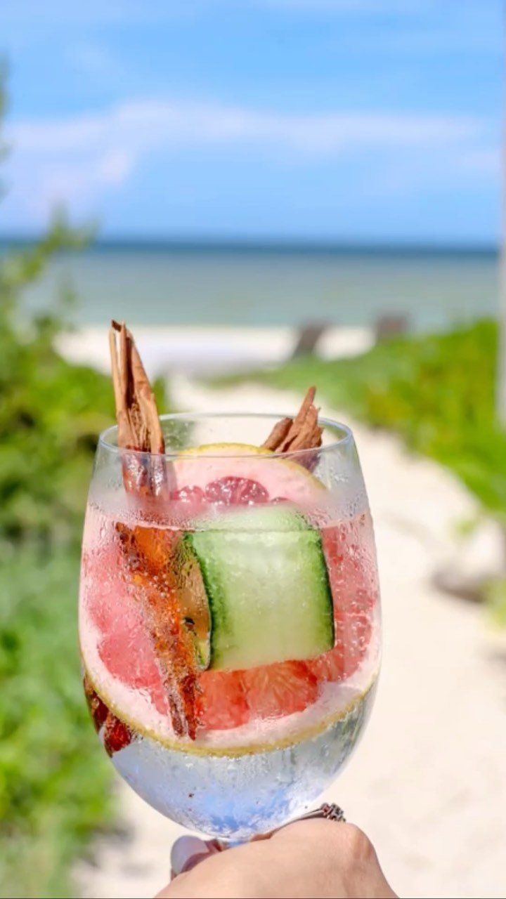 Copa de vino con un colorido cóctel de frutas, sostenida frente a una playa y un océano de fondo.