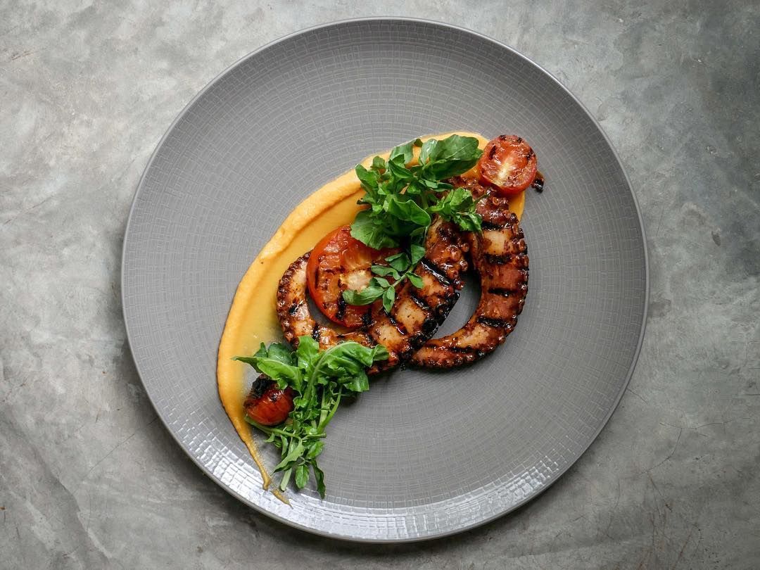 Pulpo a la parrilla con verduras en un plato gris, acompañado de una salsa cremosa.