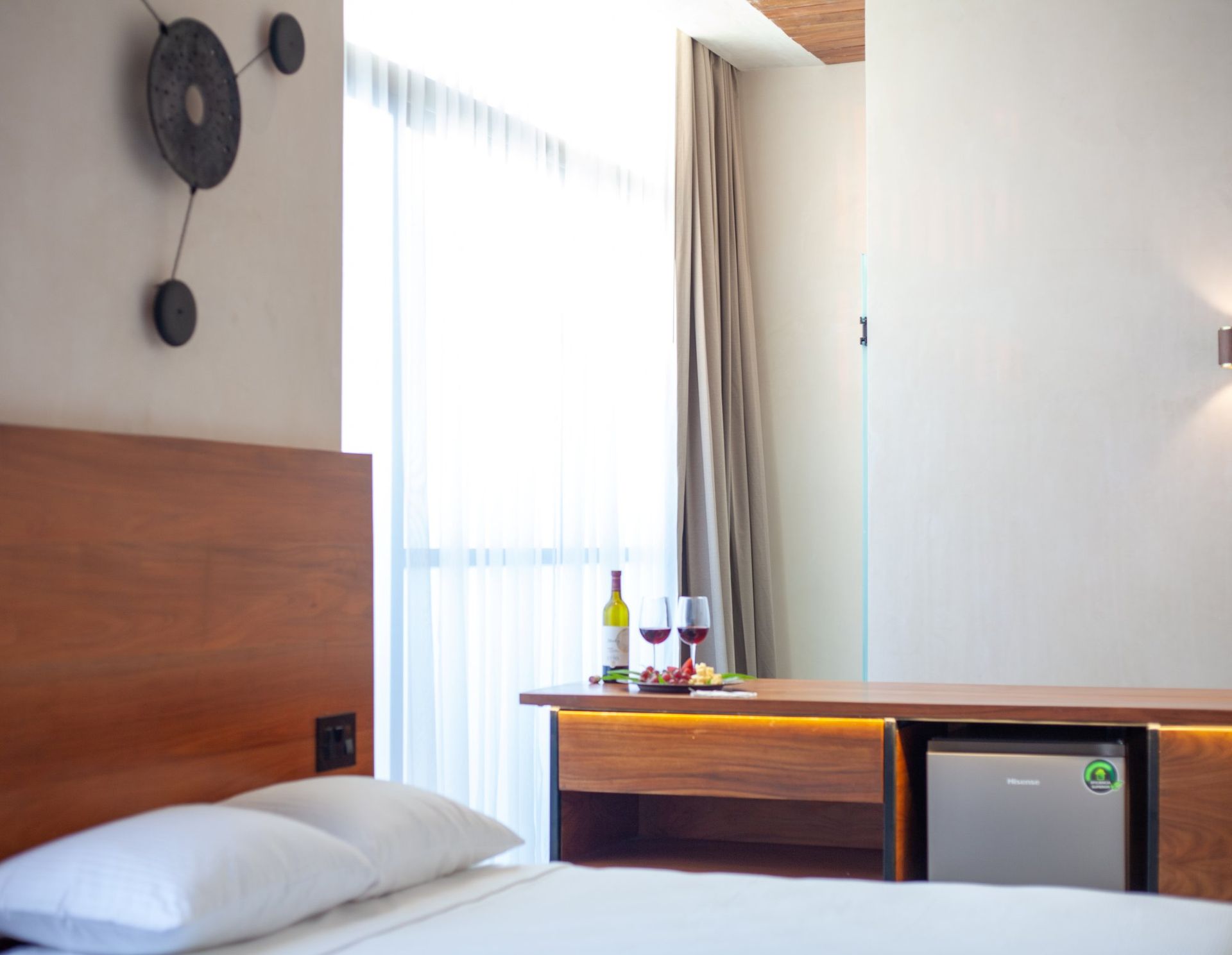Habitación de hotel moderna con cama, cabecero de madera, ventana y mininevera con bandeja en la parte superior.