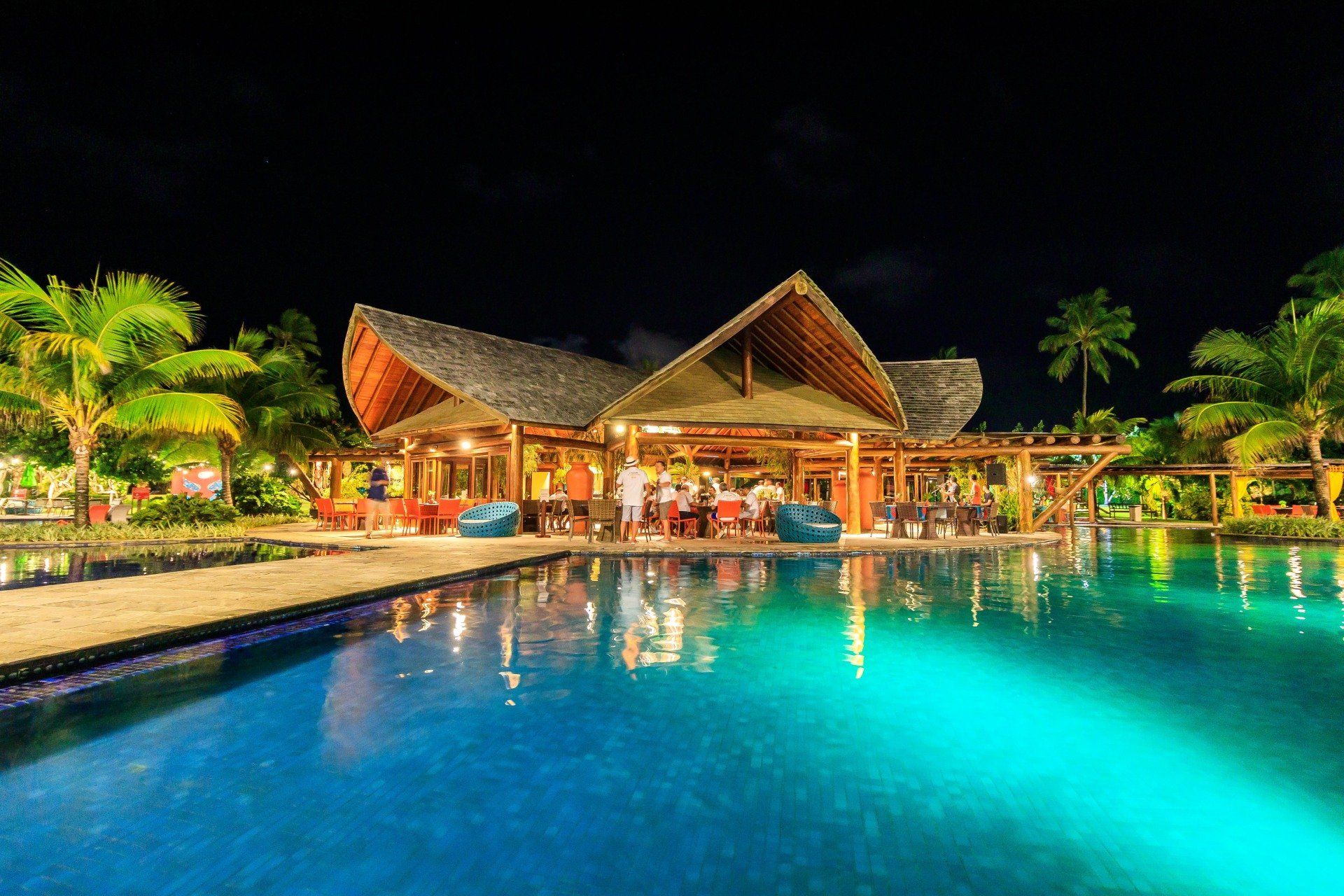 Uma grande piscina com um restaurante ao fundo à noite.