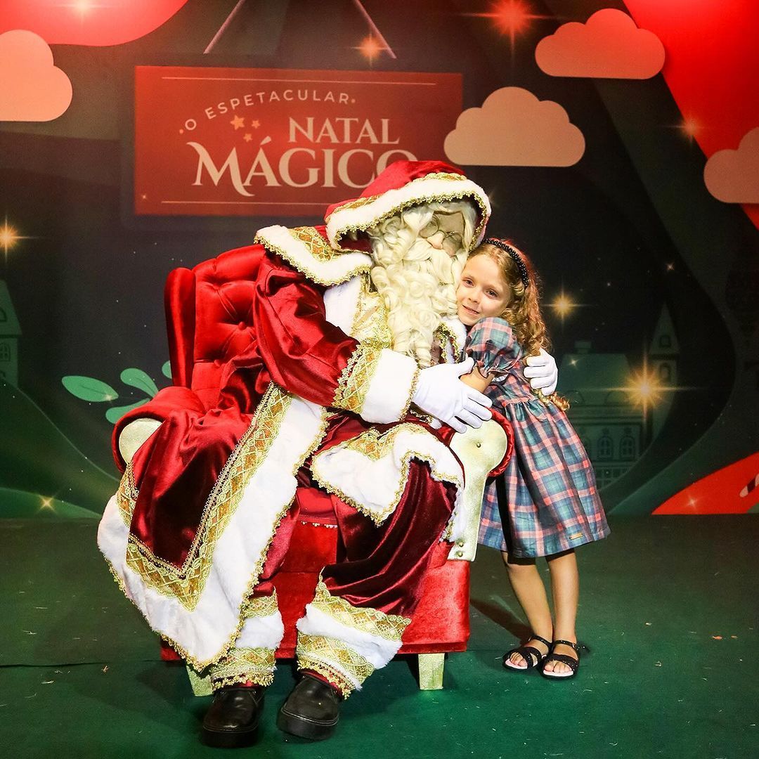 Uma menina abraça o papai noel em frente a uma placa que diz natal mágico