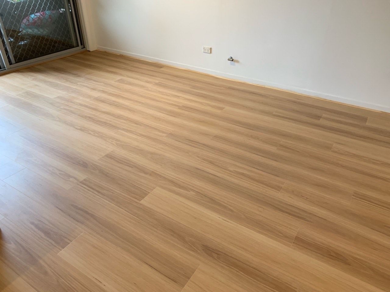 Light brown wood-look flooring in a room with a white wall and a sliding door with a screen.