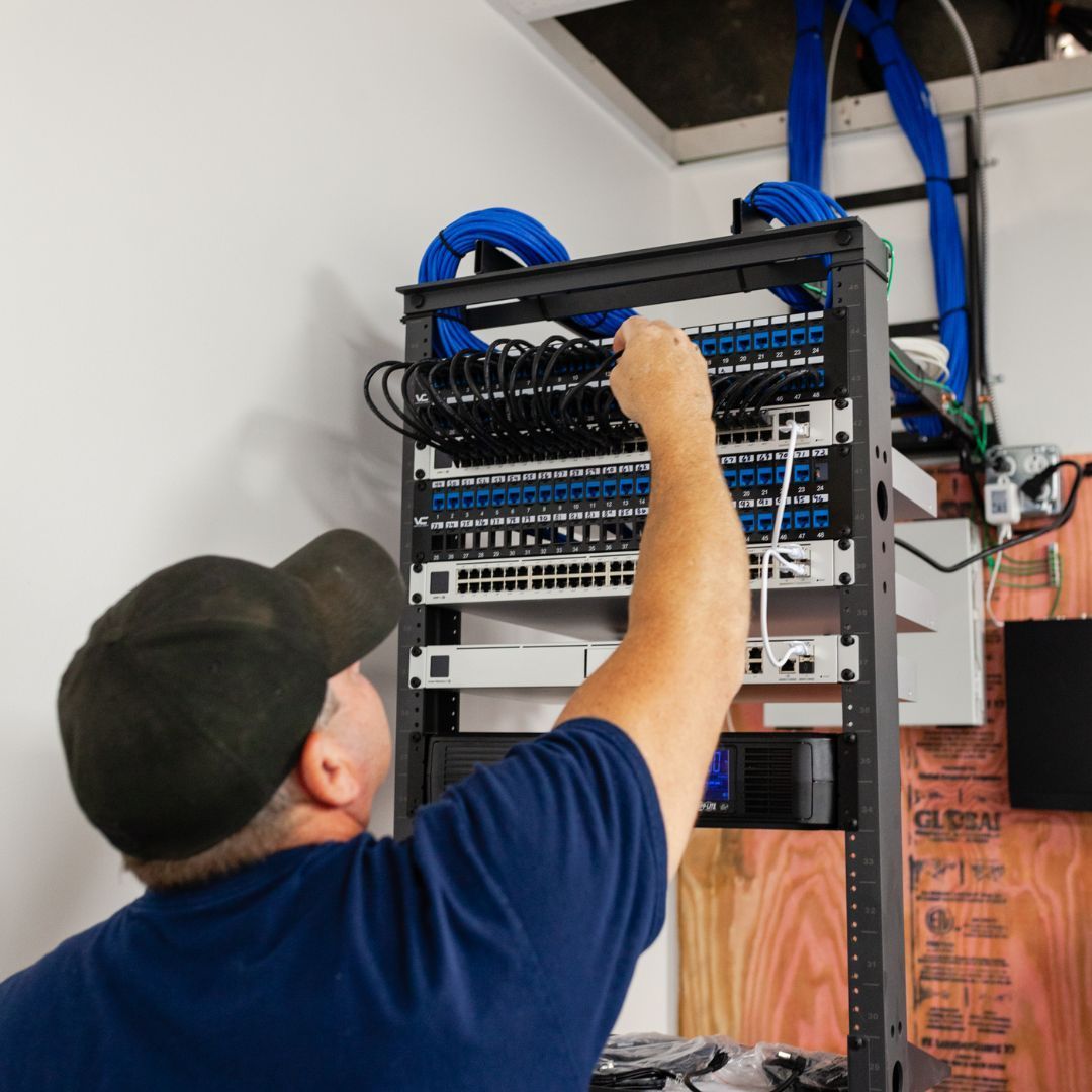 A person working on network equipment in a rack. They wear a cap and blue shirt, in a room with cables.