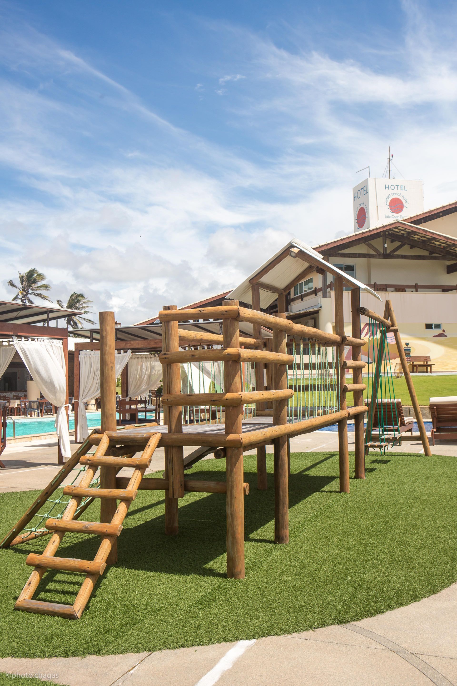 Wooden playground structure on green turf, near a building and pool, under a blue sky.