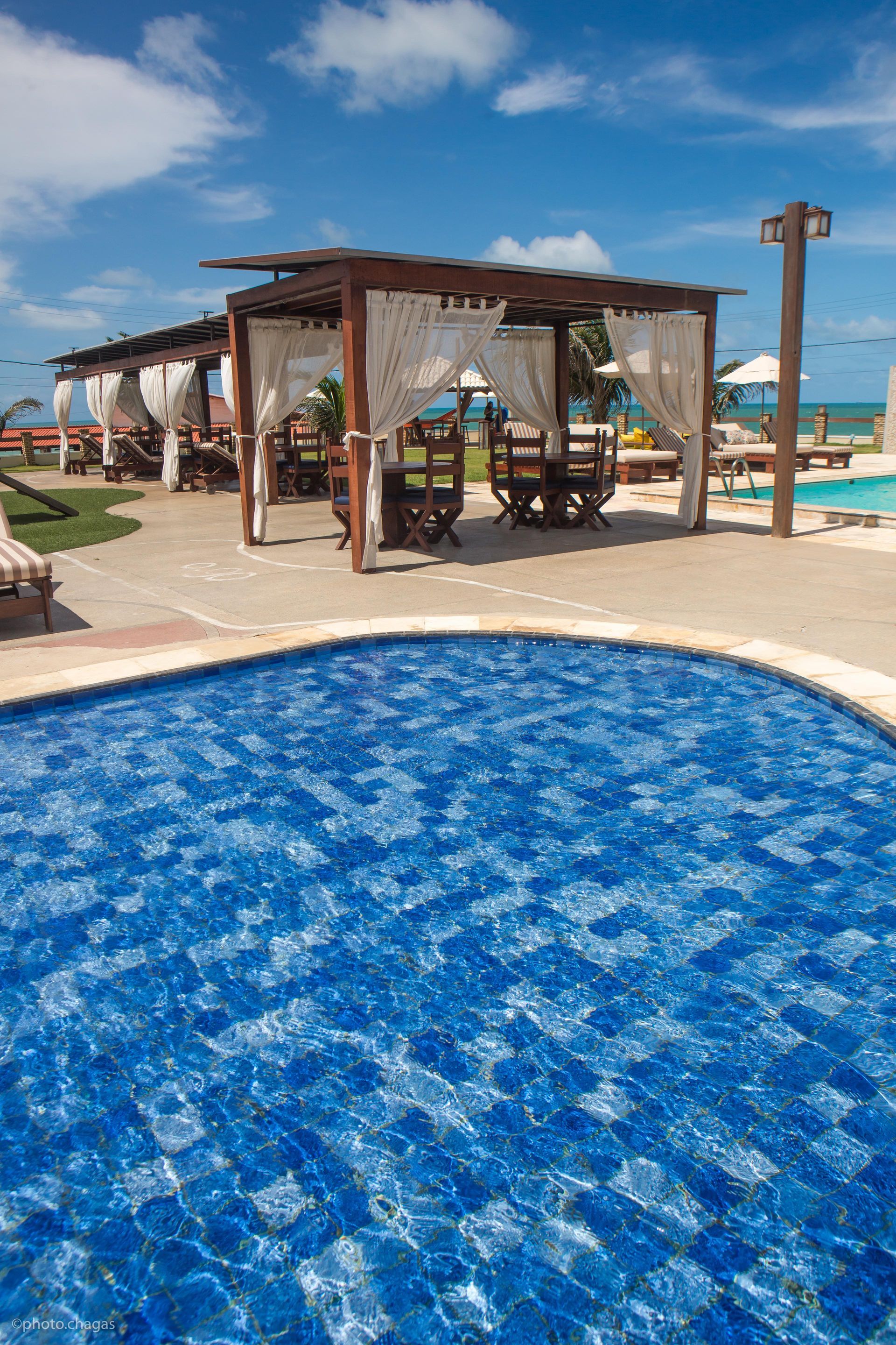 Poolside dining area with a blue-tiled pool, wooden structures, and ocean view on a sunny day.