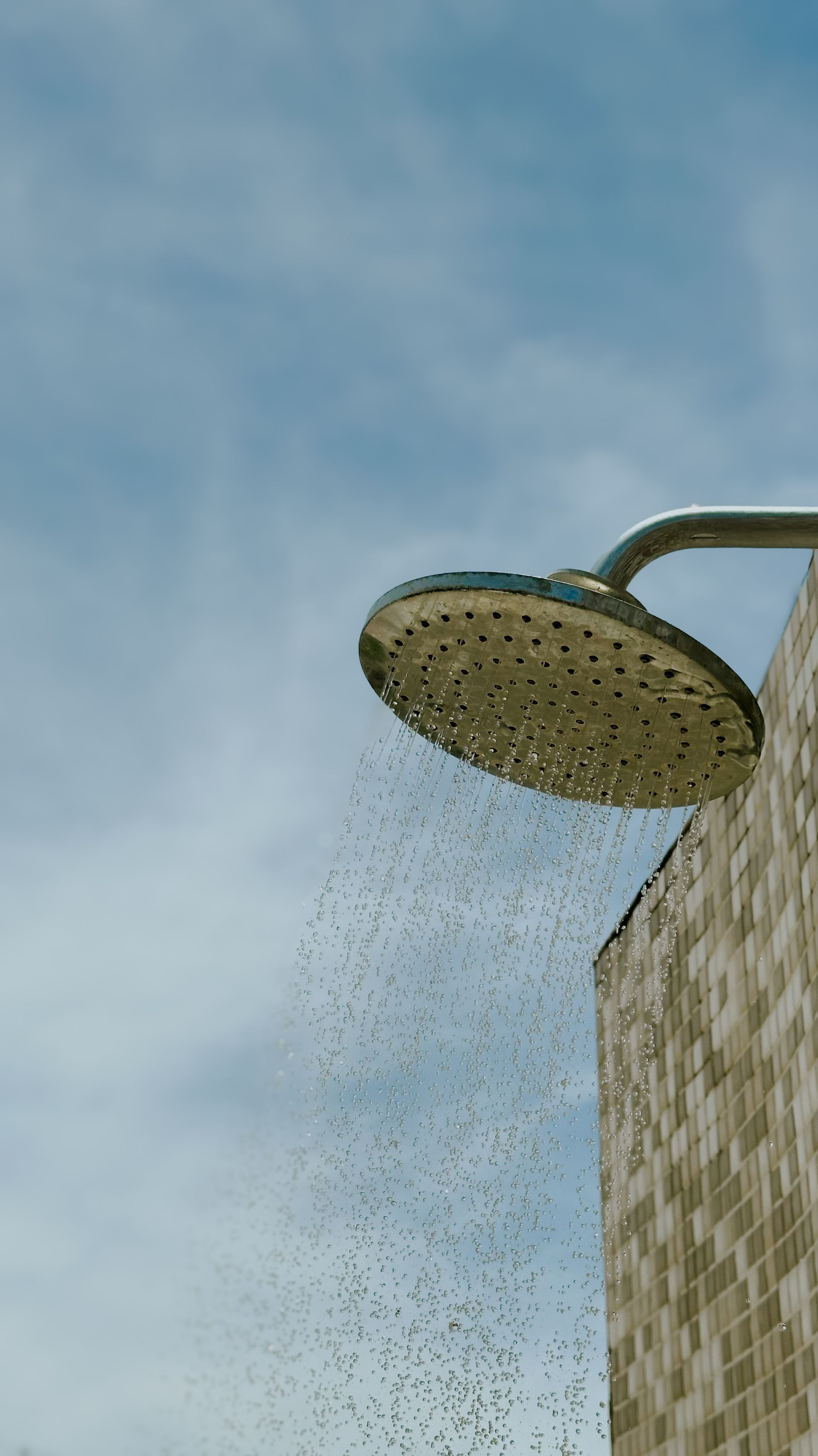 Outdoor shower head spraying water against a blue sky, mounted on a tiled wall.