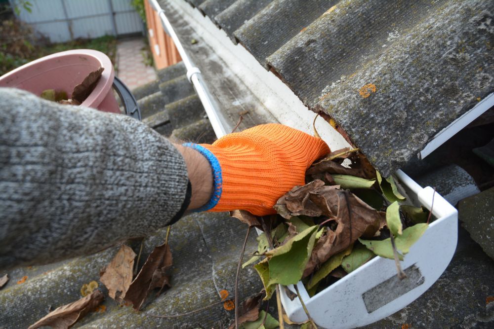 Person wearing orange glove cleaning a gutter filled with leaves.