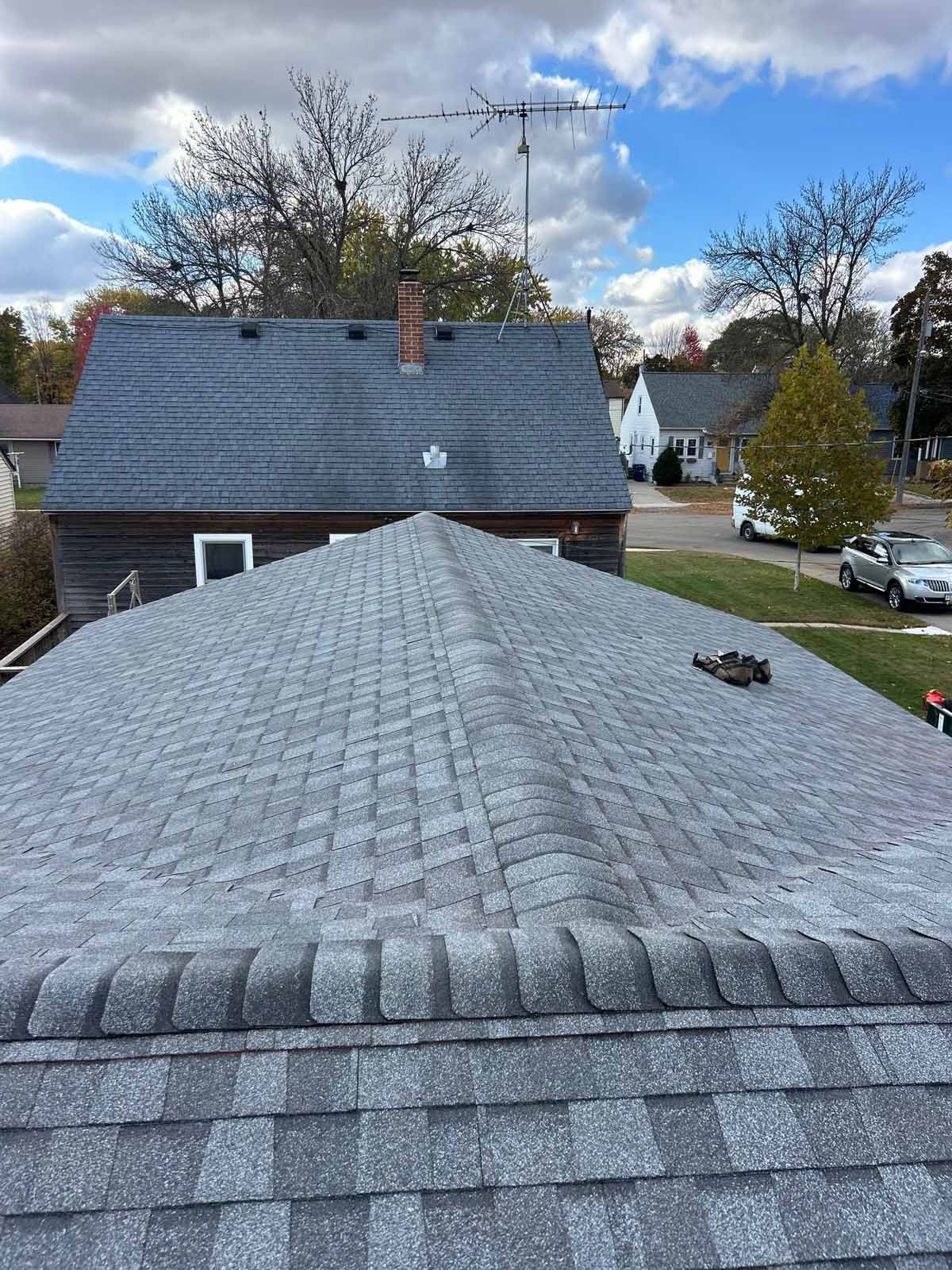Roofer working on a partially shingled roof, holding tools and wearing safety harness. Houses and trees are in the background.