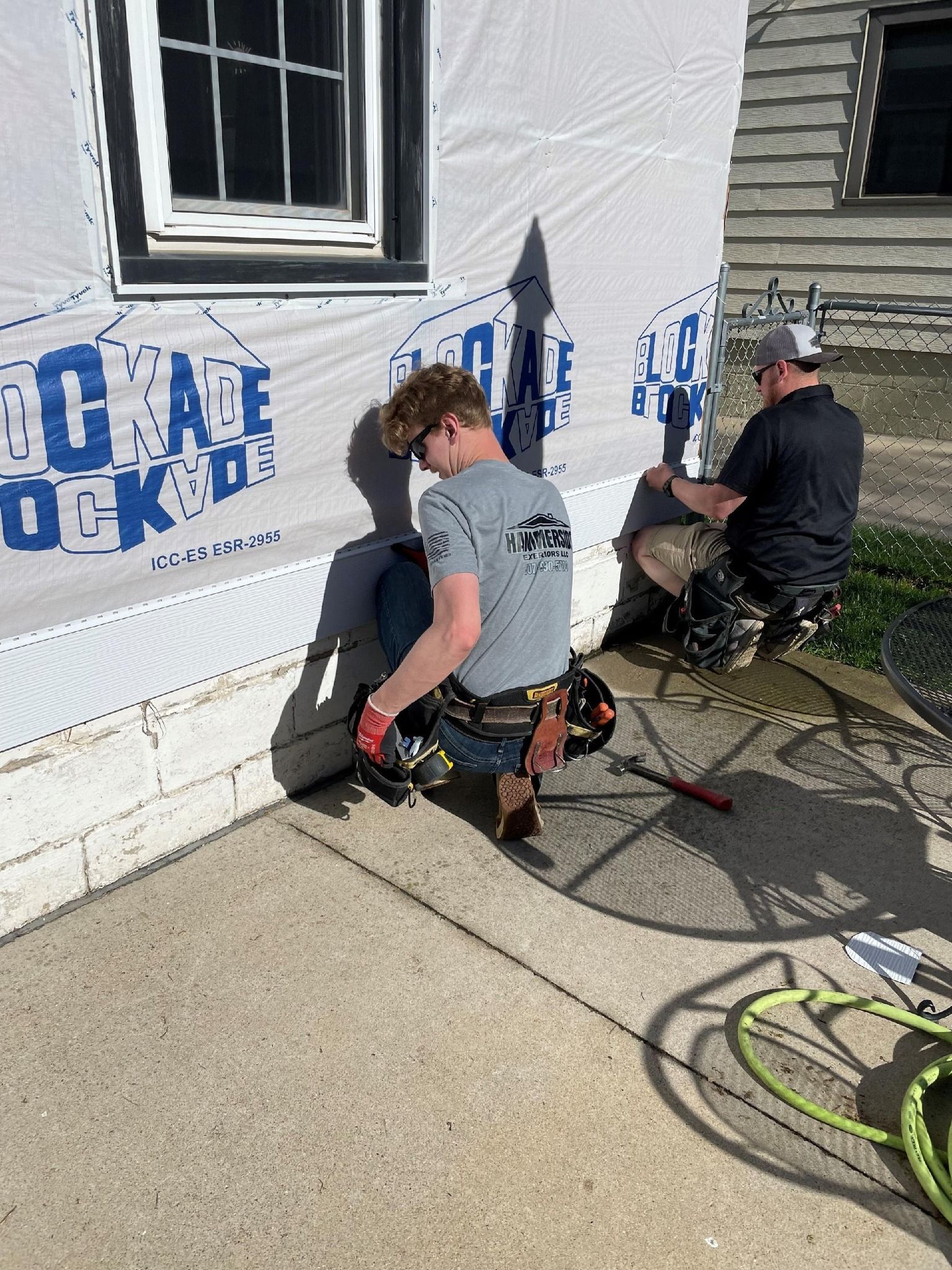 Two men working on a house exterior. One is crouched, hammering; the other is seated, holding a tool.