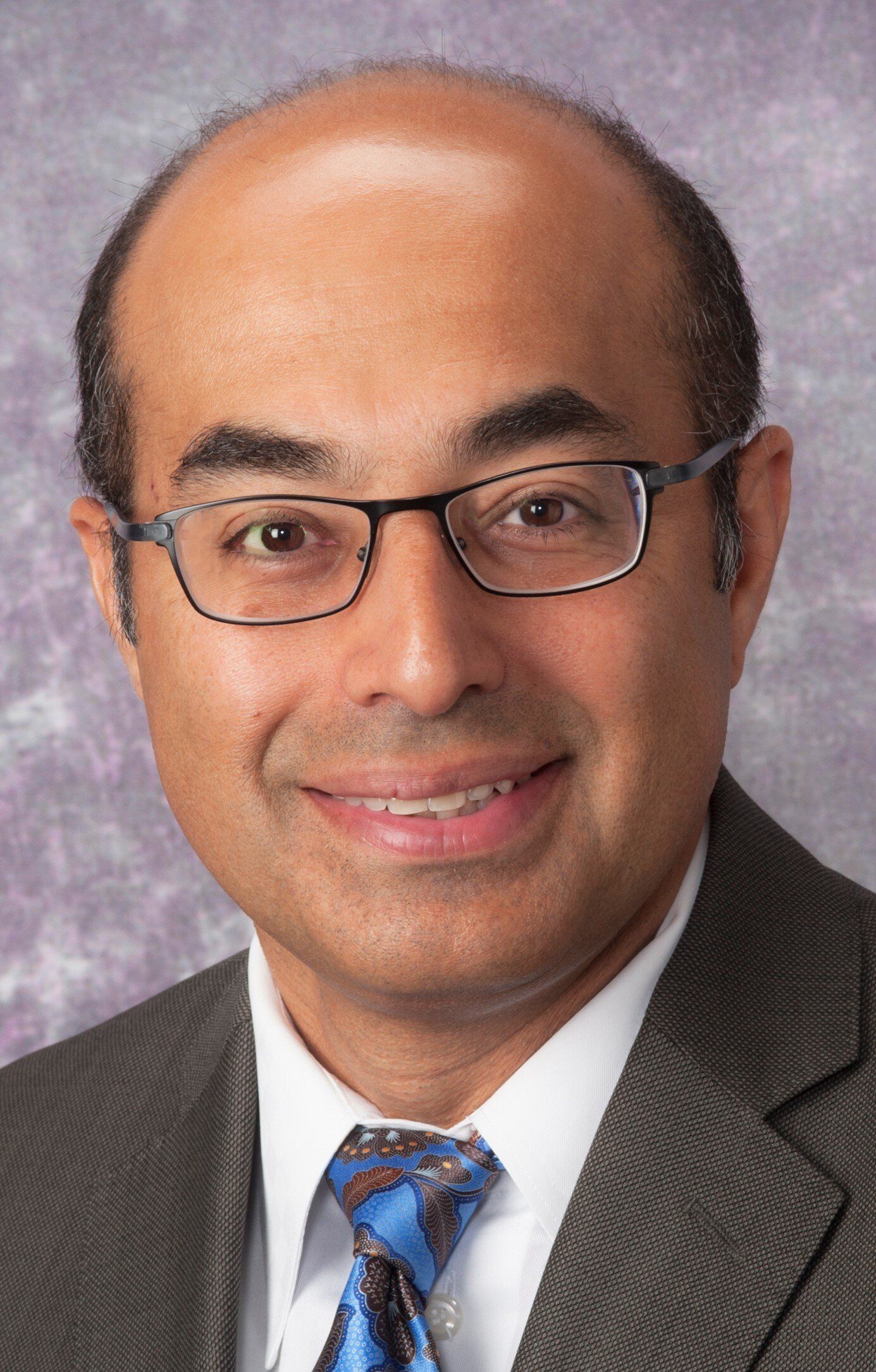 A man wearing glasses and a suit and tie is smiling for the camera.