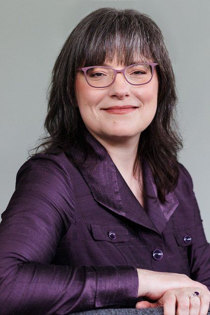 A woman wearing glasses and a purple jacket is smiling