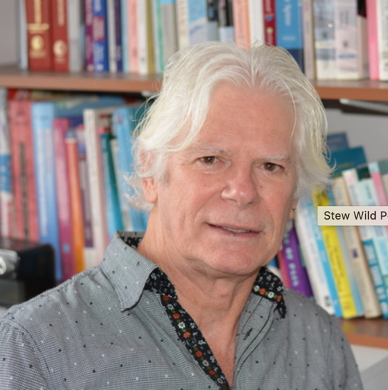 A man with white hair is standing in front of a bookshelf ..