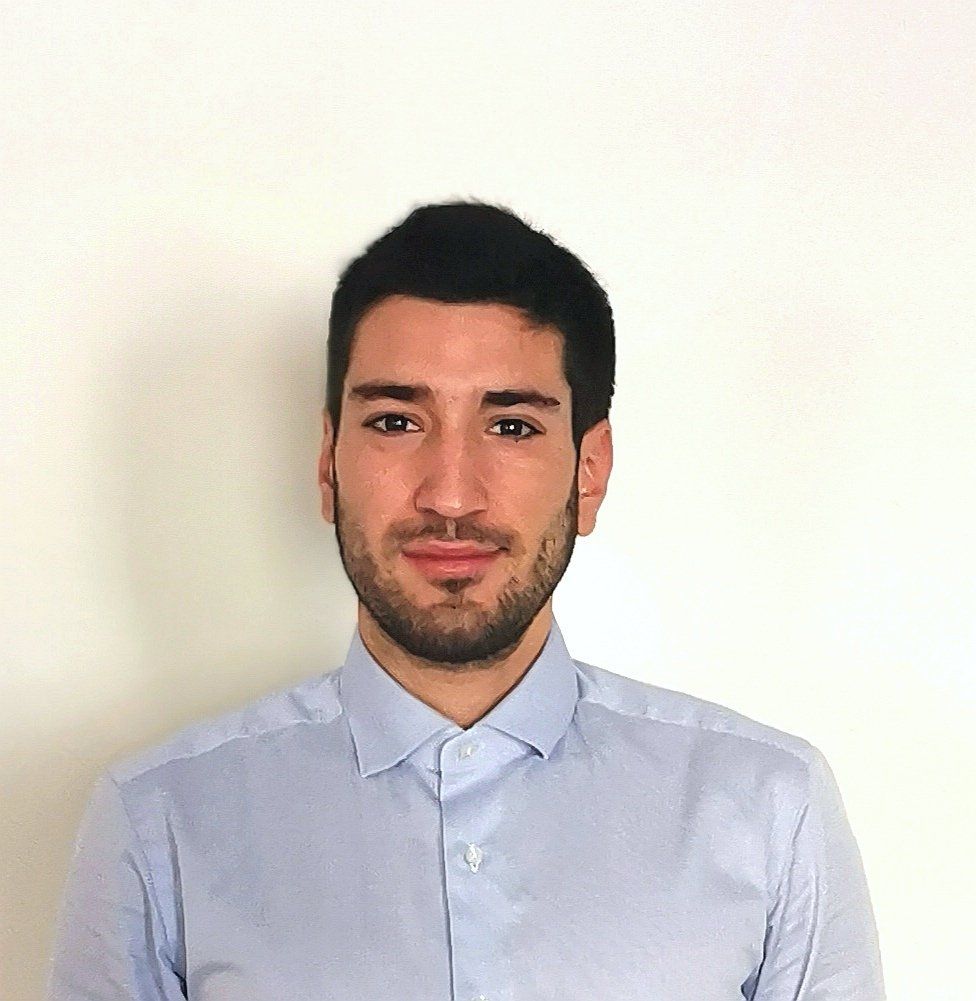 A man with a beard wearing a light blue shirt is standing in front of a white wall.