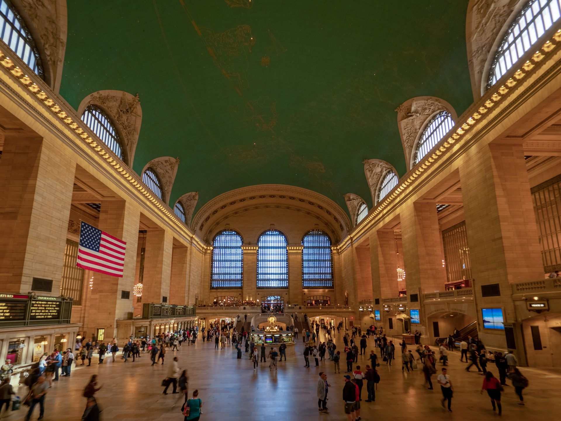 Grand Central Terminal's main concourse with a large US flag, vaulted ceiling, and commuters walking across the floor.
