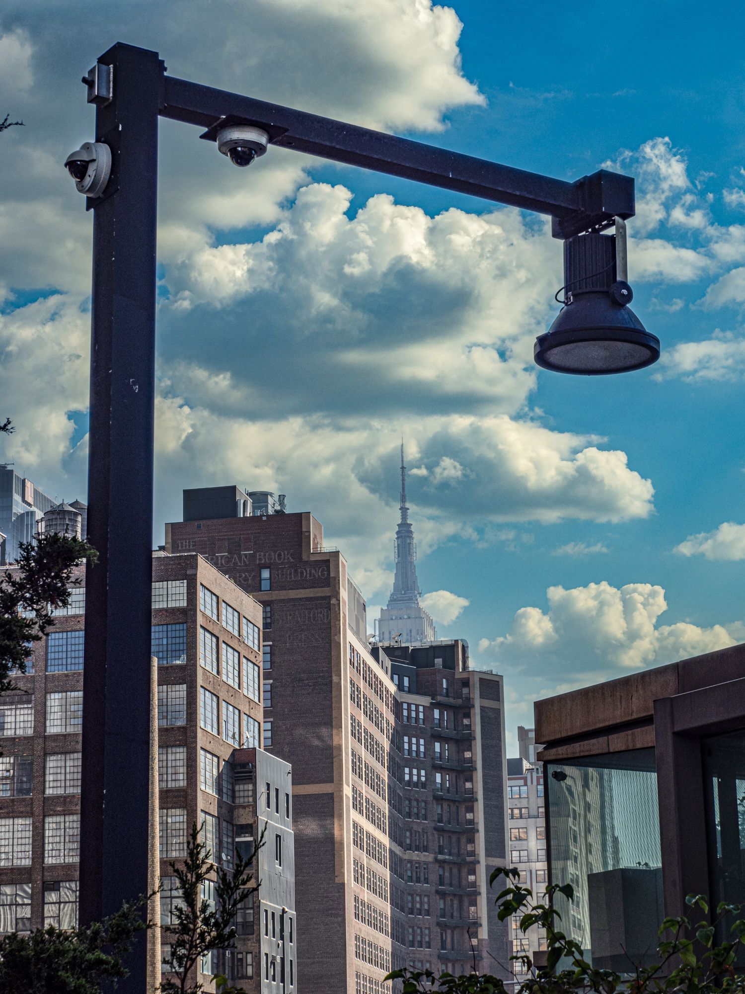 A dark metal light pole with surveillance cameras against a blue sky with clouds, framing the Empire State Building.