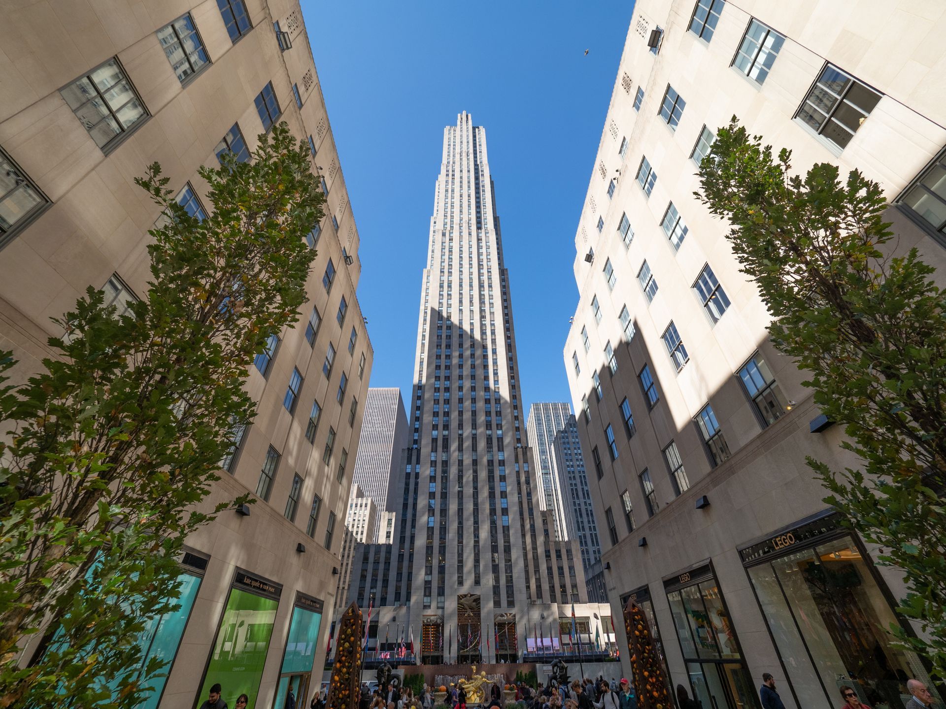 A low-angle view of 30 Rockefeller Plaza rising between two stone buildings under a clear blue sky.