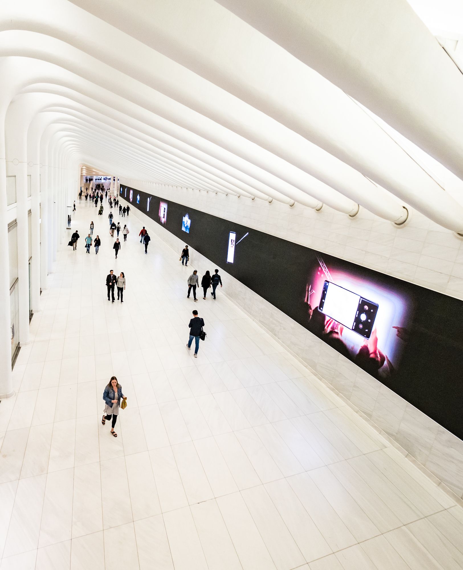 People walk through a bright, white-ribbed architectural corridor with a large digital billboard on the side wall.